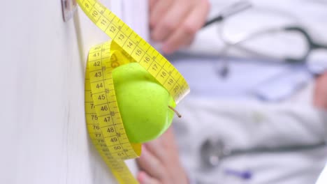 Vertical-video-of-Apple-wrapped-in-measure-tape-with-blurred-Nutritionist-taking-notes-in-Background