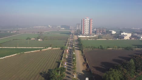 Toma-Aérea-De-Un-Dron,-La-Cámara-Del-Dron-Avanza-Hacia-Una-Carretera-Que-Atraviesa-Los-Campos-Con-Muchos-Edificios-Grandes-De-Gran-Altura-Y-Casas-Residenciales-Visibles