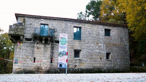 Hermosa-Construcción-Antigua-Con-Pequeñas-Ventanas-De-Las-Cuales-Cuelga-Un-Enorme-Cartel-Que-Dice-100-Años,-Rodeada-De-árboles-Y-Naturaleza.