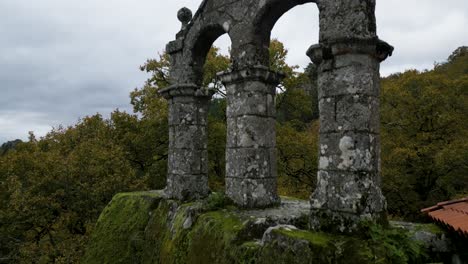 Antigua-Construcción-De-Pilares-Con-Un-Arco-Que-Sostiene-Una-Cruz-Cristiana,-Construida-Sobre-Una-Enorme-Roca-Con-Un-Corredor-Exterior-De-Un-Antiguo-Monasterio