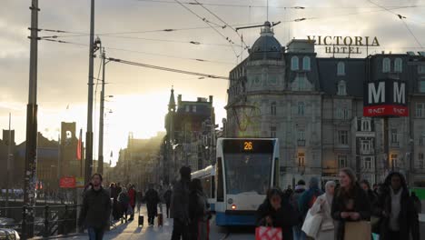 Wunderschönes-Amsterdamer-Gebäude-In-Der-Ferne,-Während-Eine-Blau-weiße-öffentliche-Straßenbahn-Während-Der-Goldenen-Stunde-Vorbeifährt-Und-Pendler-Durch-Die-Geschäftige-Stadt-In-Den-Niederlanden-Transportiert