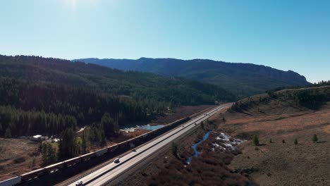 Vista-Aérea-Del-Tren-Bnsf-Que-Viaja-A-Lo-Largo-De-Una-Carretera-En-Montana