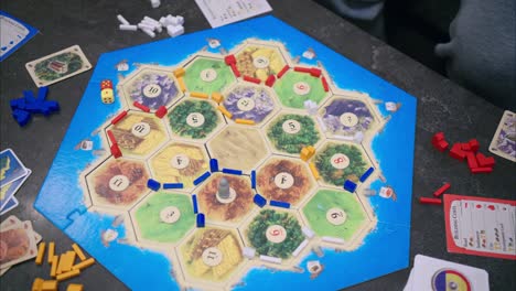 45-Grad-Draufsicht-Auf-Das-Catan-Gameplay,-Wobei-Die-Hand-Des-Spielers-Den-Räuber-Auf-Einem-Ziegelsteinplättchen-Platziert-Und-Einer-Anderen-Person-Eine-Karte-Aus-Der-Hand-Stiehlt