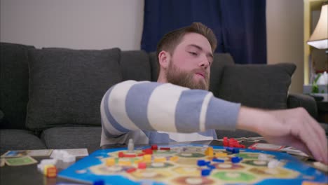 Ein-Bärtiger-Mann-Spielt-In-Seinem-Wohnzimmer-Das-Brettspiel-Catan,-Würfelt,-Zieht-Ressourcenkarten-Und-Fügt-Sie-Seiner-Hand-Hinzu