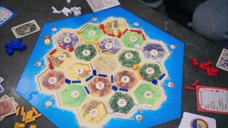 Vista-Aérea-De-45-Grados-Del-Juego-De-Catan-Con-Cuatro-Jugadores-En-Una-Mesa-De-Piedra-Con-Lanzamiento-De-Dados,-Construcción-De-Carreteras-Y-Colocación-De-Asentamientos-Con-La-Mano-Del-Jugador-Azul.