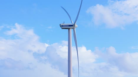 Vista-Lateral-De-Grandes-Palas-De-Turbina-De-Molino-De-Viento-Girando-Contra-El-Cielo-Azul-Con-Nubes-Blancas