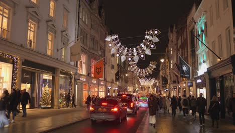 Toma-De-Autos-Y-Peatones-Moviéndose-Por-Una-Calle-Iluminada-Con-Adornos-Navideños-Por-La-Noche