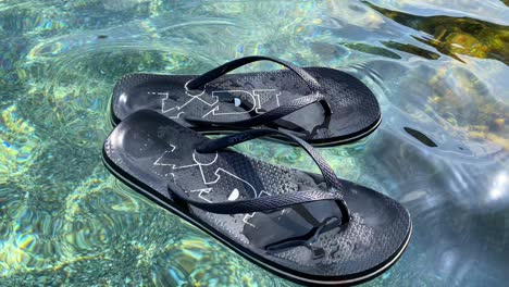 Chanclas-Flotantes-De-Supermercado-Lidl-Negro-En-Agua-De-Mar-Turquesa-Transparente-Junto-A-La-Playa-En-España,-Divertidos-Y-Soleados-Zapatos-De-Vacaciones-De-Verano,-Toma-De-4k