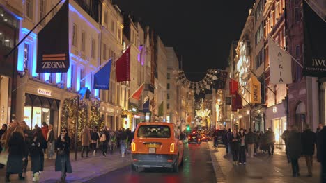 Toma-De-Autos-Y-Peatones-Caminando-Por-Una-Calle-Iluminada-Llena-De-Adornos-Navideños-Por-La-Noche