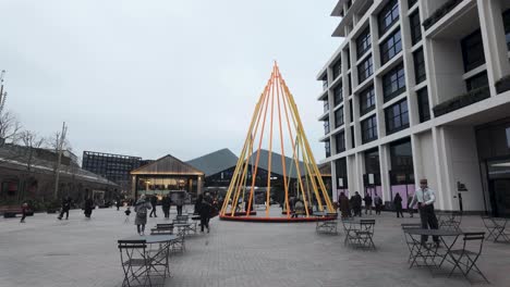 Illuminated-Artwork-At-Lewis-Cubitt-Square,-Coal-Drops-Yard,-Kings-Cross-With-People-Walking-Past-On-Overcast-Day