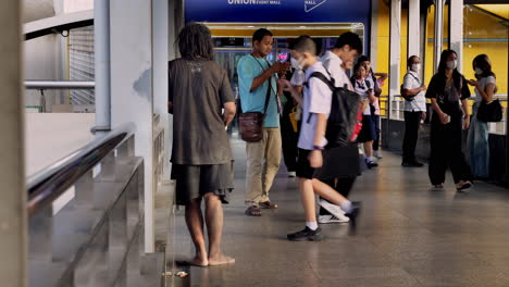 A-homeless-man-is-begging-as-pedestrians-are-passing-by-at-a-footbridge-crossing-in-front-of-the-Union-Mall-entrance-in-Bangkok,-Thailand