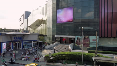 Shoppers-going-in-and-out-of-a-shopping-center-while-some-are-passing-by-and-buying-some-street-food-from-vendors-selling-in-front-of-a-mall-in-Bangkok,-Thailand