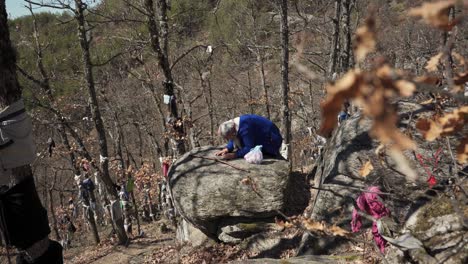 An-old-woman-praying-and-doing-an-ancient-ritual-in-front-of-a-sacred-natural-stone-arch-at-Skribina-Sanctuary,-known-for-its-healing-powers-at-the-foot-of-Western-Rhodope-Mountains-in-Bulgaria