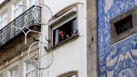 Anciana-Chismeando-A-Través-De-Una-Ventana-Rodeada-De-Palomas-En-Portugal