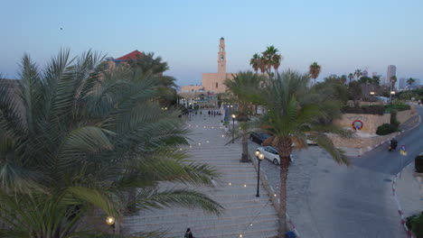 Ciudad-Antigua-De-Jaffa-Al-Atardecer-Con-Muchas-Familias-Visitando-Restaurantes,-Tiendas-Y-Bares-En-El-Puerto:-Empuje-Con-Drones-Entre-Los-árboles-De-Dátiles