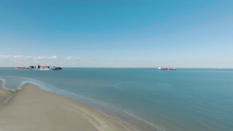 Una-Vista-Aérea-Captura-Un-Buque-Portacontenedores-Cargado-Que-Pasa-Por-El-Extremo-Este-De-La-Isla-De-Galveston-Mientras-Una-Pareja-Camina-Por-La-Playa-Bajo-Un-Cielo-Azul-Claro-En-Galveston,-Texas.