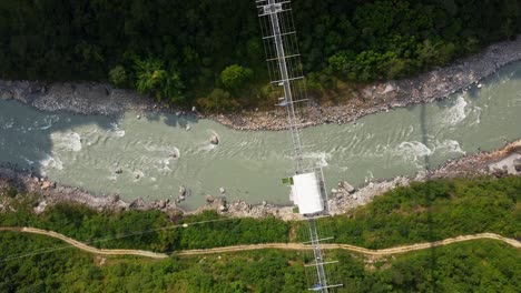 Toma-Aérea-De-Arriba-Hacia-Abajo-Del-Río-Y-El-Puente-Colgante-En-Nepal-Durante-El-Día-Soleado