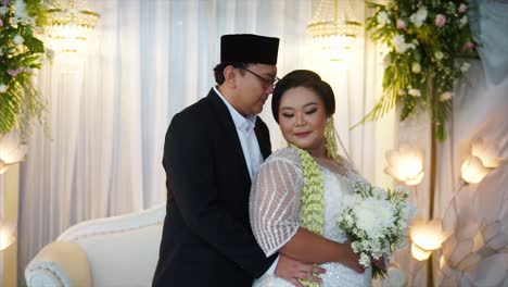 Groom-hugs-the-bride-from-the-back-at-an-Indonesian-wedding-party,-they-wear-traditional-Indonesian-wedding-dresses-and-carry-a-bouquet