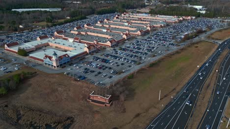 Toma-Aérea-Amplia-Que-Muestra-Un-área-De-Estacionamiento-Abarrotada-En-Un-Centro-Comercial-Y-Un-Vehículo-Conduciendo-En-Una-Autopista-En-Estados-Unidos