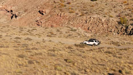 Toma-De-Seguimiento-De-Un-Todoterreno-Blanco-Conduciendo-Por-Un-Camino-De-Tierra-A-Través-Del-Paisaje-Del-Desierto-De-Utah-En-Un-Día-Soleado