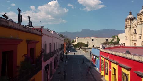 Vuelo-Aéreo-Entre-Casas-Coloridas-Con-Gente-Caminando-En-Oaxaca-De-Juárez-Y-La-Catedral-Al-Fondo