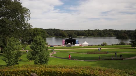 Familias-Y-Niños-En-Trentham-Gardens,-Escenario-Junto-Al-Lago-Preparado-Para-Conciertos.