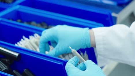 Over-the-shoulder-shot-of-a-lab-technician-in-lab-jacket-and-blue-medical-gloves-on-the-assembly-line-packing-cannabis-pre-rolls-into-a-tube-that-will-be-sold-in-dispensaries