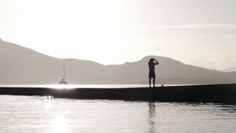 La-Silueta-De-Un-Hombre-Da-La-Bienvenida-Al-Amanecer-En-Un-Muelle,-Abrazando-El-Calor-Del-Sol-En-Meditación