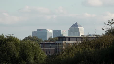 In-Zeitlupe-Ziehen-Wolken-In-Der-Ferne-Hinter-Einem-Wohngebäude-Am-Finanzviertel-Canary-Wharf-Vorbei,-Während-Züge-Zwischen-Zwei-Baumgruppen-Hindurchfahren