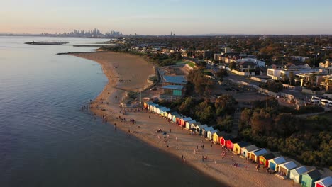 Vídeo-De-Drones-Sobrevolando-Las-Famosas-Cajas-De-Baño-De-Brighton,-Con-El-Hermoso-Horizonte-De-La-Ciudad-De-Melbourne-En-El-Horizonte-Lejano-Al-Atardecer