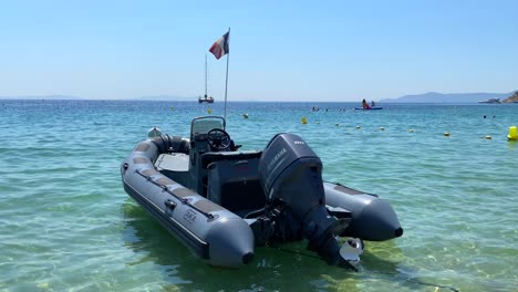 Lancha-Inflable-A-Motor-Con-Bandera-Francesa-Flotando-En-Agua-De-Mar-Turquesa-Transparente-En-Un-Día-Soleado-En-Cavalière-Lavandou-Al-Sur-De-Francia,-Divertidas-Vacaciones-En-La-Playa,-Toma-Estática-De-4k