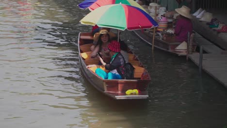 Establishing-Shot,-Man-is-rowing-a-boat-with-tourist-in-Thailand-Floating-Market,-Assorted-Hats-are-displayed-on-the-boat-of-a-vendor-in-the-background