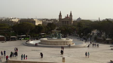 Gente-Caminando-Alrededor-De-La-Fuente-Del-Tritón-Junto-A-La-Puerta-De-La-Ciudad-De-La-Valeta,-Con-La-Iglesia-De-San-Publio-Al-Fondo.