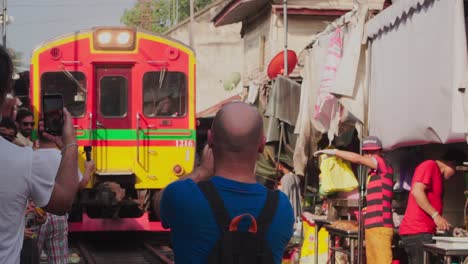 Plano-Medio,-Un-Hombre-Con-Camisa-Azul-Toma-Una-Foto-Del-Tren-Que-Se-Acerca-En-El-Mercado-Ferroviario-De-Maeklong-Samut,-Tailandia