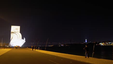 Pescadores-Y-Gente-Caminando-Frente-Al-Monumento-Padrão-Dos-Descobrimentos-En-La-Noche,-Lisboa,-Portugal