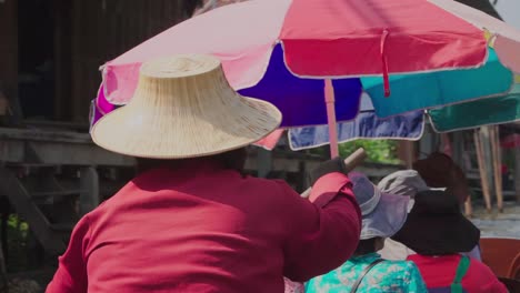 Medium-shot,-Women-in-red-with-a-straw-hat-rowing-a-tourist-boat,-scenic-view-of-Thailand-Floating-Market