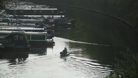 Young-man-kayaking-on-the-canal-near-boats-docked-at-the-wharf,-wide-shot