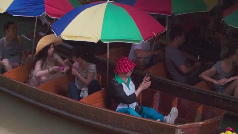 Establishing-Shot,-Wooden-boat-with-colorful-umbrella,-tourist-takes-photos-in-Thailand-floating-Market,-Tourist-Boat-Park-near-the-market-in-the-background