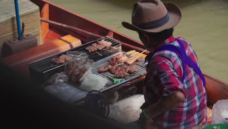 Plano-Medio,-Hombre-Con-Sombrero-De-Vaquero,-Remando-En-Un-Bote-Vendiendo-Barbacoa-En-El-Mercado-Flotante-De-Tailandia