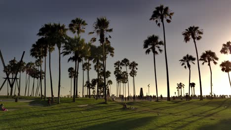 Gente-Caminando-Por-El-Paseo-Marítimo-De-Venice-Beach-Durante-La-Hora-Dorada-Del-Atardecer-Bajo-Palmeras-Durante-La-Pandemia-De-Covid-19---Toma-Panorámica