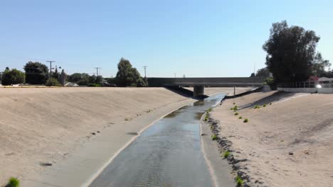 Aerial-view-over-people-walking-towards-a-bridge,-at-the-LA-River,-on-a-sunny-day,-in-Los-Angeles,-California,-USA---Low,-dolly,-drone-shot