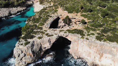 Ein-Drohnenschuss,-Der-Rückwärts-Von-Einem-Meeresbogen-Wegfliegt,-Mit-Ein-Paar-Touristen,-Die-Das-Bild-In-Der-Nähe-Von-Cala-Varques-Auf-Mallorca-Betreten