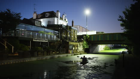 Hombre-Haciendo-Kayak-En-Un-Canal-Verde-Rodeado-De-Casas-Por-La-Noche,-Plano-Amplio