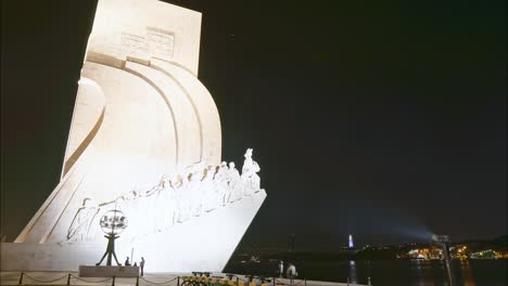 Timelapse-from-the-illuminated-Padrão-dos-Descobrimentos-monument-at-night-with-the-25-de-Abril-Bridge-and-Sanctuary-of-Christ-the-King-in-the-background,-Lisbon,-Portugal