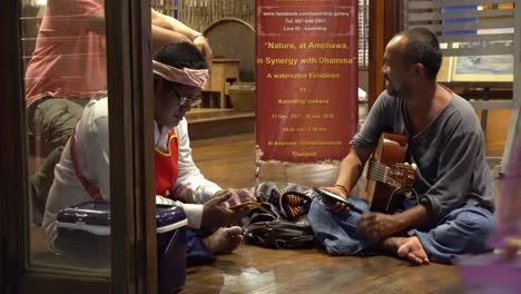 Thai-Guitarist-Musician-Sitting-on-Floor-Before-Performance-and-Looking-at-Smartphone