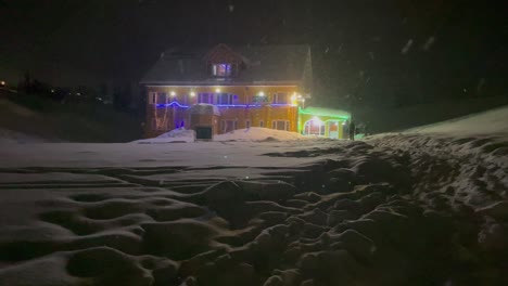 Gente-Caminando-Por-La-Noche-En-Un-Paisaje-Nevado-Fuera-De-La-Casa-Adornada-Con-Luces-Navideñas-En-Medio-De-Las-Nevadas-En-Invierno-En-Gulmarg,-India