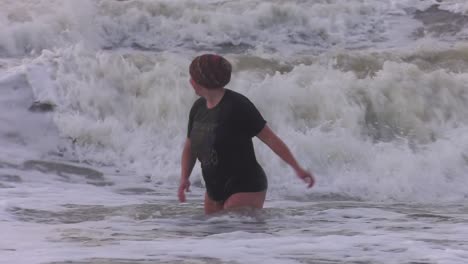 Clumsy-Dutch-woman,-stumbling-on-the-cold,-foaming-sea-at-New-Year's-dive-in-Texel-Beach-in-North-Holland,-Netherlands---Medium-Tracking-shot