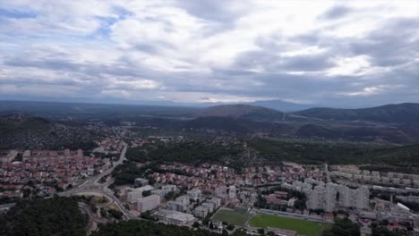 Drohnenschuss-Flog-Rückwärts-über-Die-Stadt-Sibenik-Und-Die-Berge-Im-Hintergrund-Unter-Bewölktem-Himmel