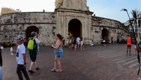 Hombres-Y-Mujeres,-Turistas-Caminan-Y-Toman-Fotografías-Con-Sus-Cámaras-Cerca-De-Una-Antigua-Pared,-Arco-Y-Reloj-De-Una-Plaza-En-El-Casco-Antiguo-De-Cartagena-De-Indias,-Colombia