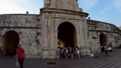 Un-Grupo-De-Turistas-Y-Un-Hombre-Con-Camisa-Roja-Caminan-Cerca-De-Un-Antiguo-Arco-O-Puerta-De-Piedras-Que-Ingresa-A-Una-Plaza-Del-Casco-Antiguo-De-Cartagena-De-Indias,-Colombia